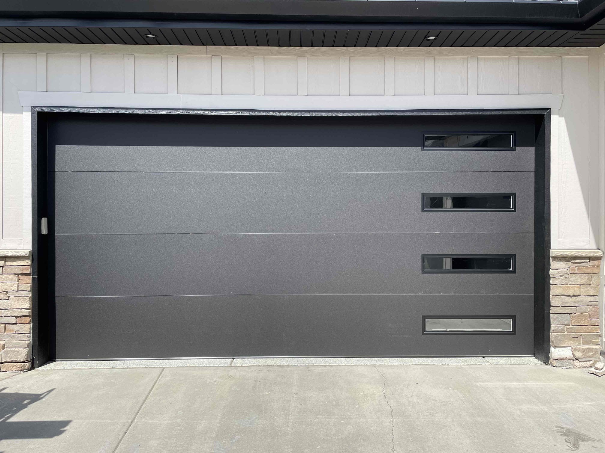 Sleek modern garage door in matte black with four narrow horizontal windows aligned vertically on the right side, framed by white siding and stone accents on either side.