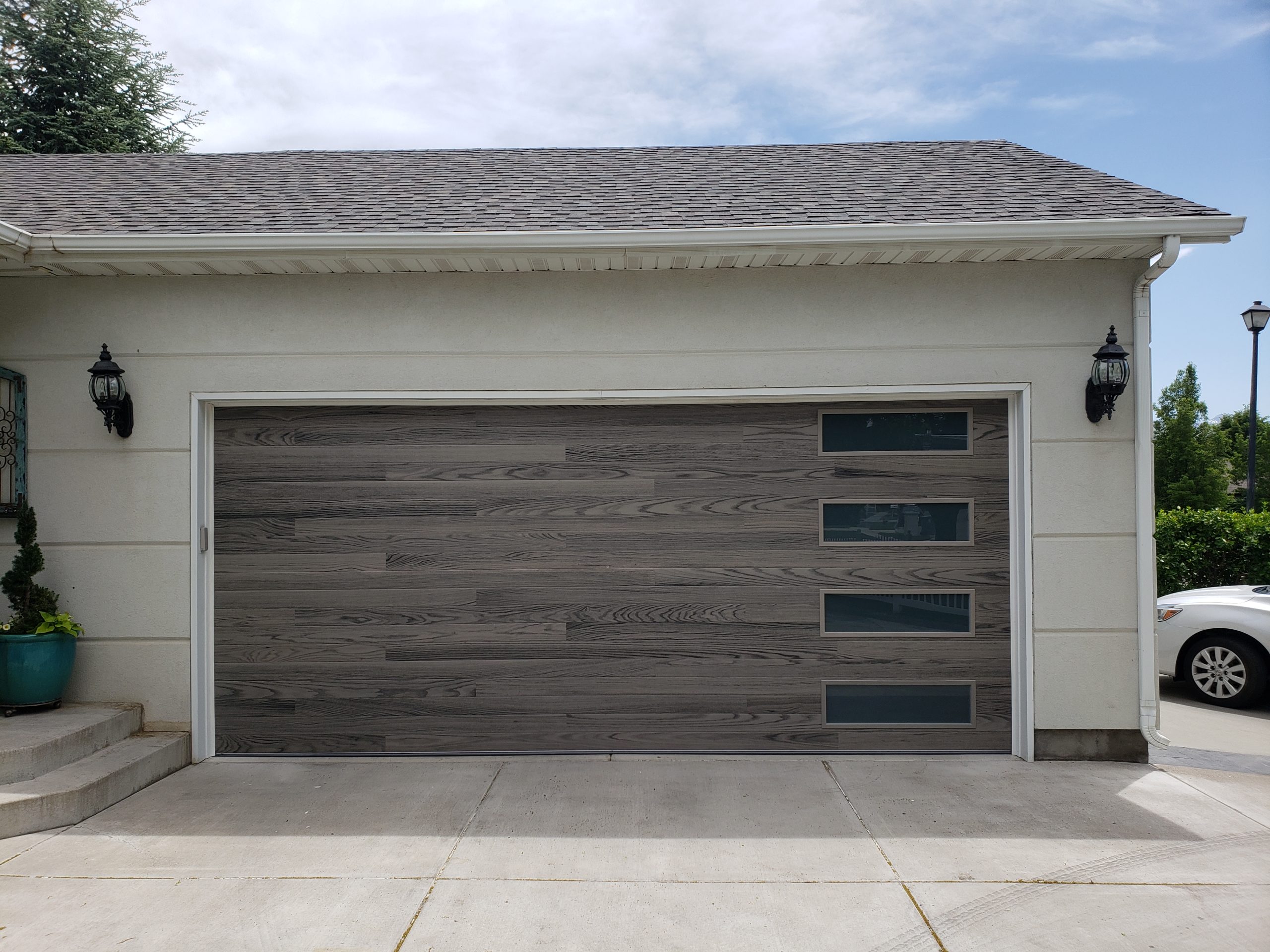 Modern horizontal plank garage door in a gray wood finish with four narrow rectangular windows aligned vertically on the right side, set on a cream stucco home with decorative black sconces.