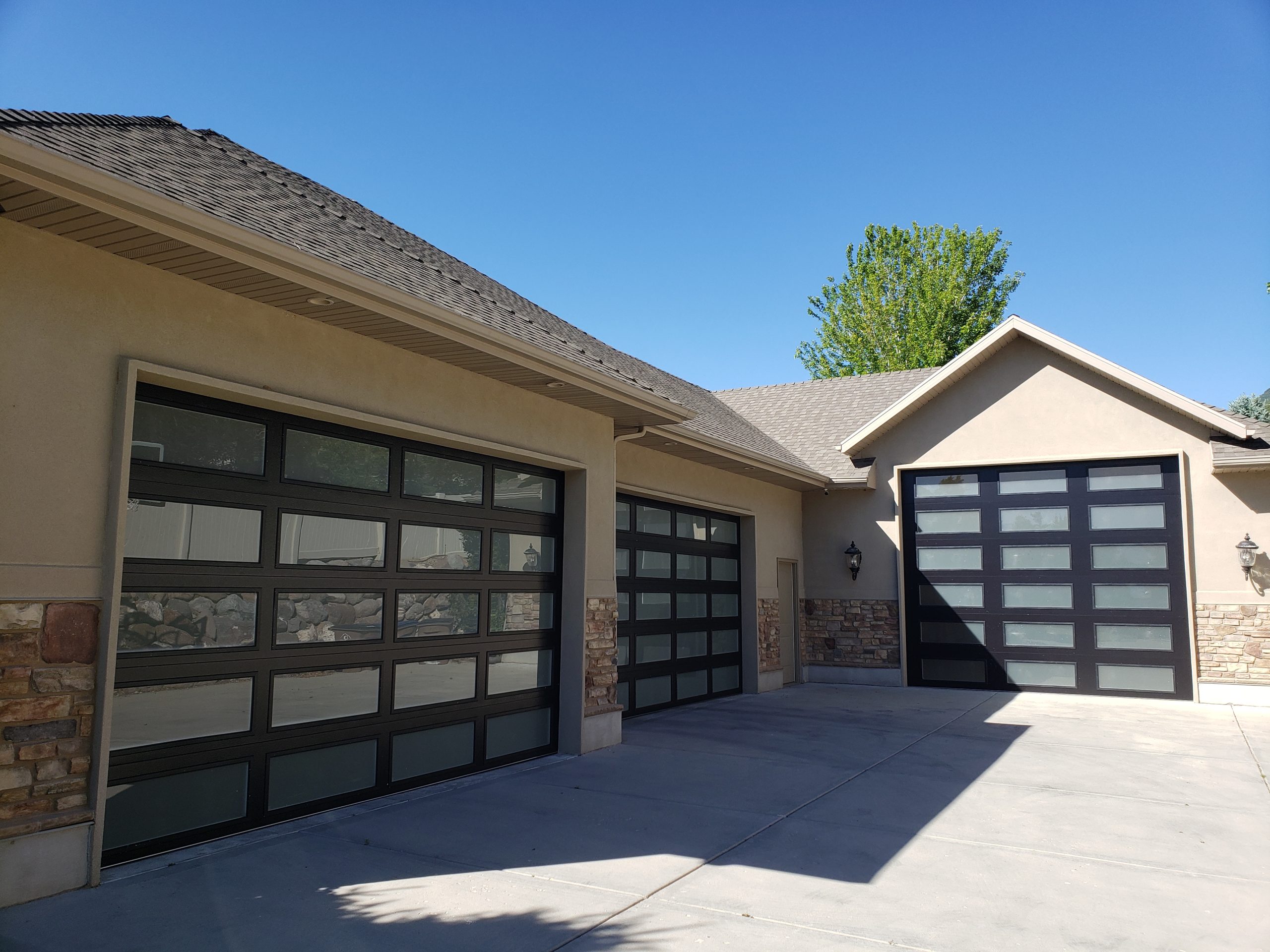Modern home with three black-framed frosted glass garage doors, set against a beige exterior with stone accents and a concrete driveway under a clear blue sky.