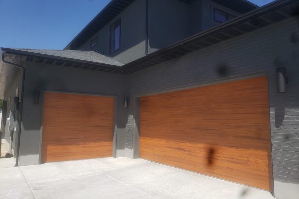 Modern home with two side-by-side garage doors featuring a warm horizontal wood finish, set against a dark gray exterior with minimalist black sconces and a clean concrete driveway.