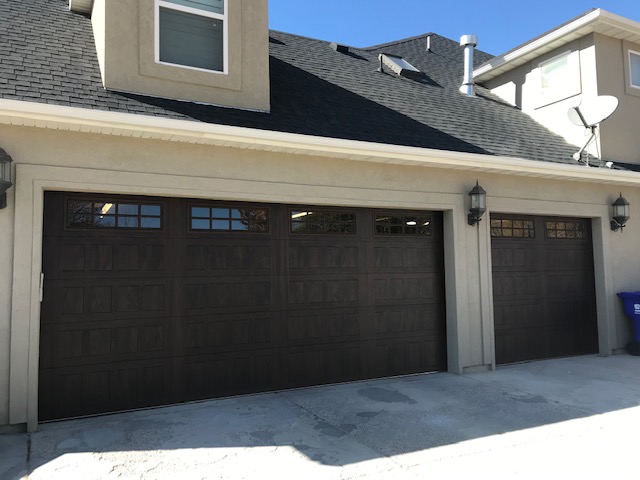Three-section brown garage doors with decorative window panels installed on a beige stucco home with a pitched roof and dormer window, under a clear blue sky.