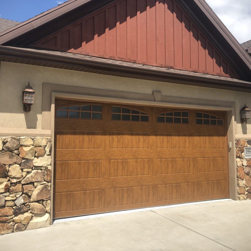 Wood-look garage door with arched window panels on a home featuring stone siding accents, beige stucco, and a red gable roof under a partly cloudy sky.