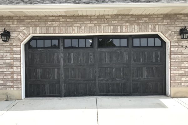 Dark wood-look double garage door with upper rectangular window panels on a light brick home exterior, featuring a clean concrete driveway and black wall-mounted lanterns