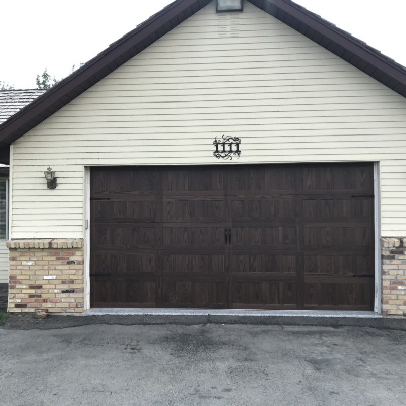 Traditional garage with a dark wood-look door on a cream-colored house featuring horizontal siding and a brick wainscoting base, with wall-mounted lantern-style lights on either side.