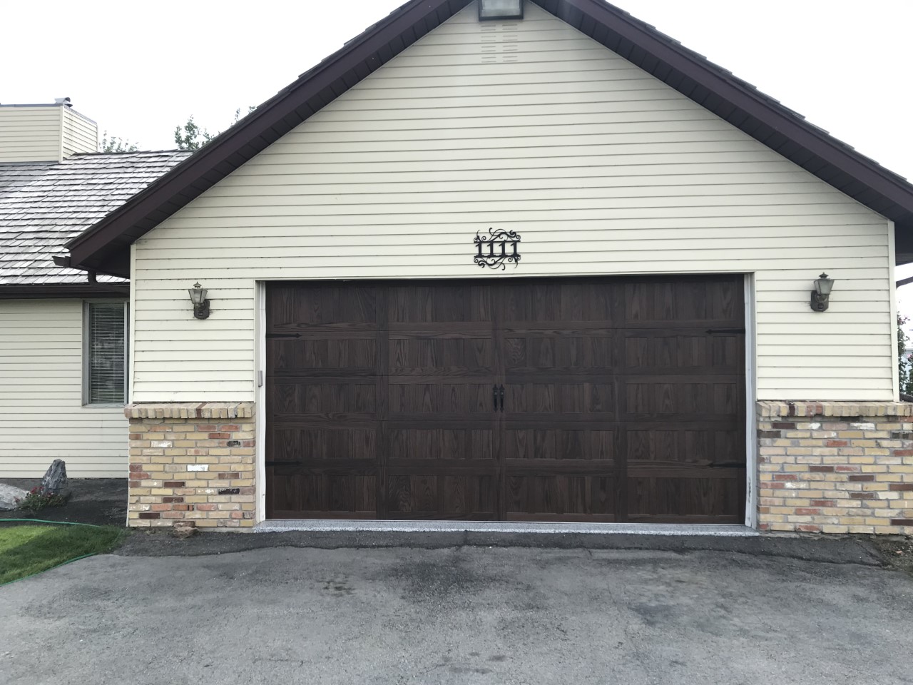 Traditional garage with a dark wood-look door on a cream-colored house featuring horizontal siding and a brick wainscoting base, with wall-mounted lantern-style lights on either side.