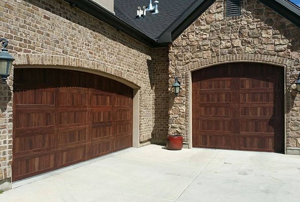 Elegant brick and stone home with two arched wooden garage doors featuring a classic raised-panel design, set beneath a steep roofline and accented with decorative wall sconces.