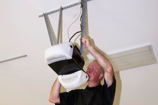Man installing a ceiling-mounted garage door opener, working with wiring and metal brackets on a garage ceiling during a mechanical setup or repair.