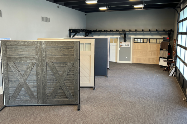 Garage door showroom with multiple freestanding door panel samples in various styles and finishes, including rustic wood, modern, and carriage-style designs, displayed on a carpeted floor under bright lighting.