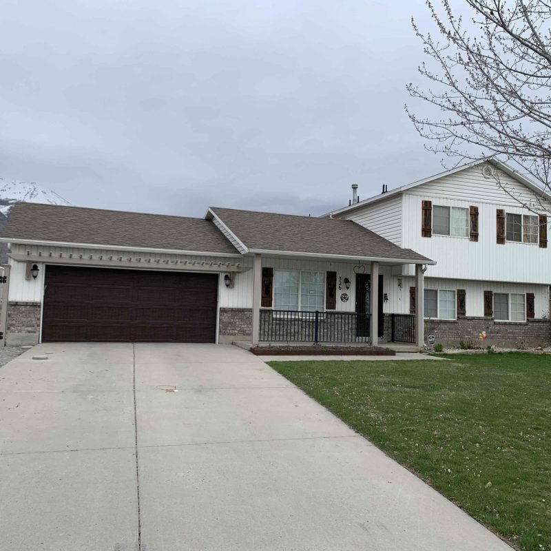 Split-level suburban home with a dark brown garage door, white siding, stone wainscoting, and a wide concrete driveway, set against a cloudy sky with a snow-capped mountain partially visible in the background.