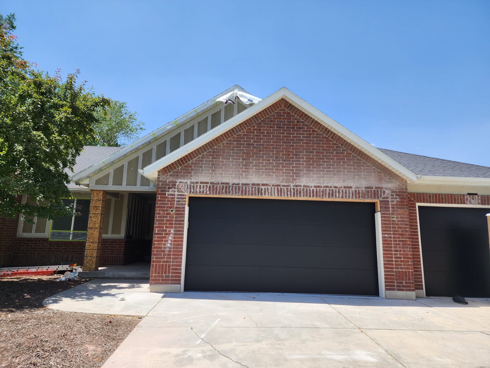 Red brick suburban home with modern matte black garage doors, steep gabled roof, and a clean concrete driveway under a bright blue sky.