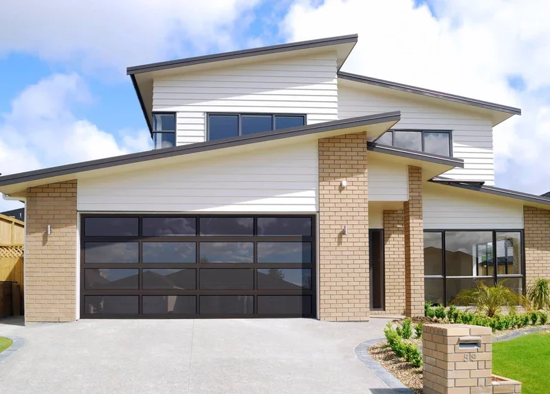 Contemporary two-story home with a large glass-panel garage door, light brick and white siding exterior, sloped rooflines, and a curved driveway leading to a landscaped front yard.