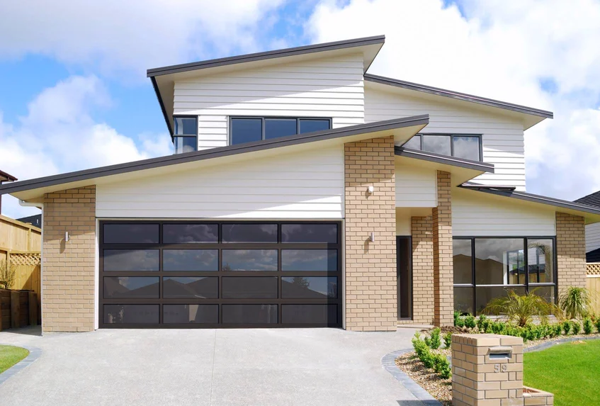Contemporary two-story home with a large glass-panel garage door, light brick and white siding exterior, sloped rooflines, and a curved driveway leading to a landscaped front yard.