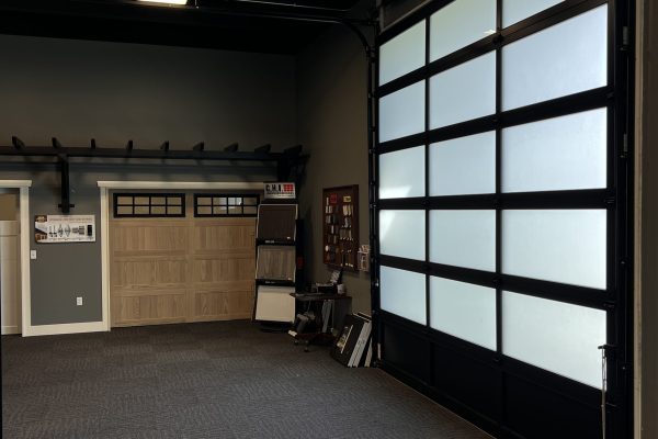 Spacious garage door showroom featuring a large frosted glass-panel garage door on the right and multiple garage door style samples on the back wall under ceiling lights.