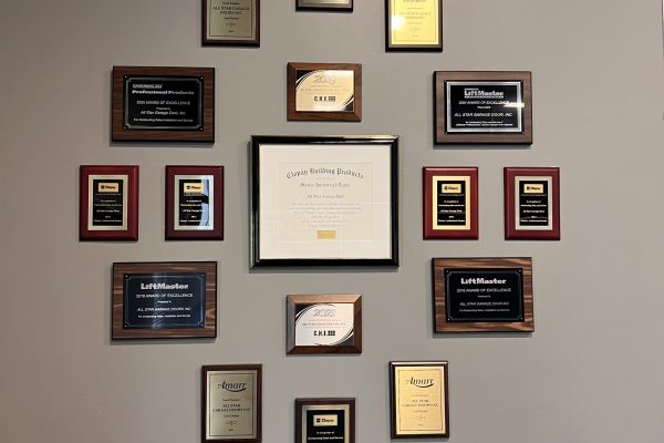 Wall-mounted display of numerous awards and certificates arranged in a symmetrical grid, featuring various plaques and a central framed achievement on a gray background.