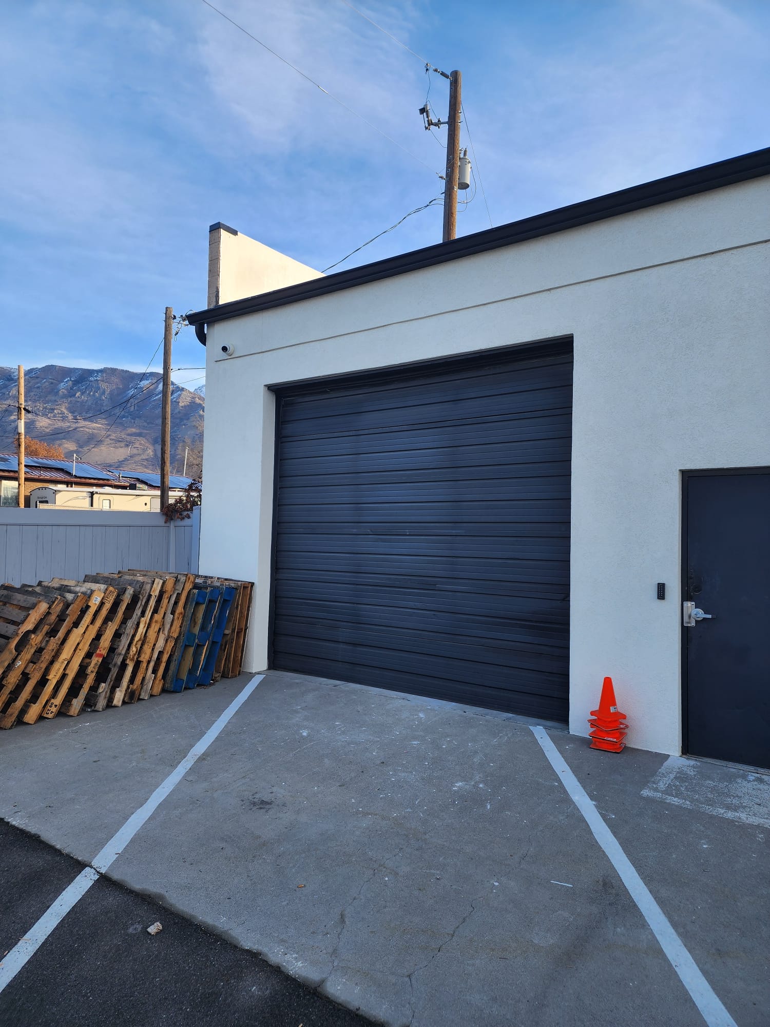 Modern industrial building with a large black roll-up garage door, adjacent side entry door, and concrete driveway lined with parking stripes, wooden pallets, and an orange safety cone.