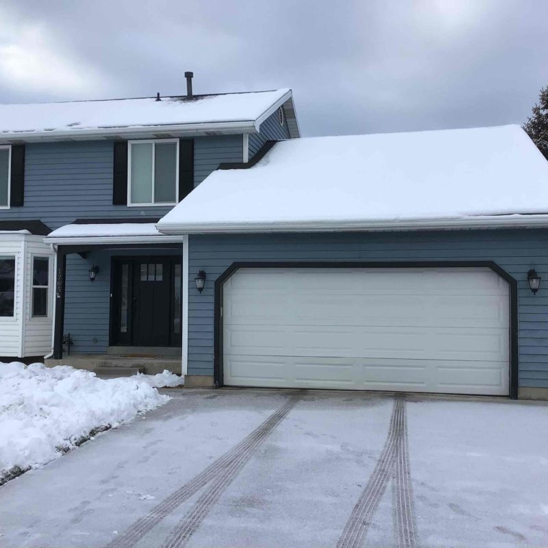 Two-story blue house with white trim and a white paneled garage door, partially covered in snow with visible tire tracks on the driveway under a cloudy winter sky.