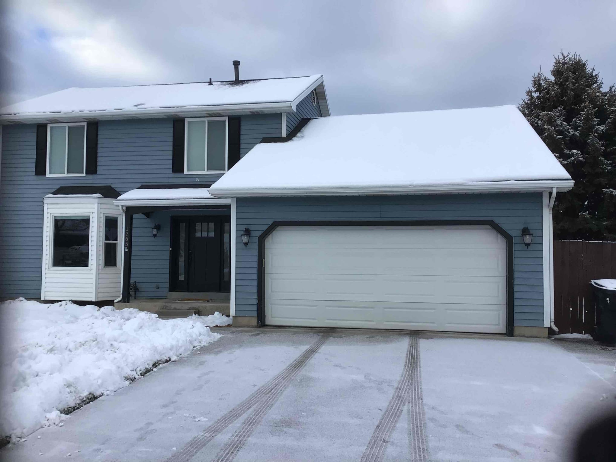 Two-story blue house with white trim and a white paneled garage door, partially covered in snow with visible tire tracks on the driveway under a cloudy winter sky.