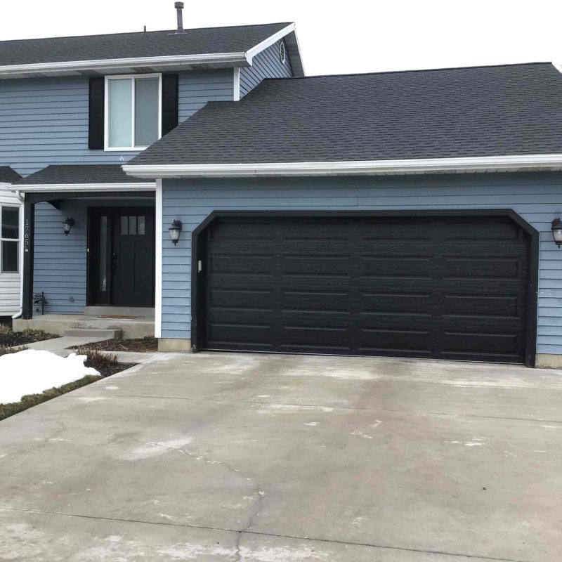 Two-story home with blue horizontal siding and a black raised-panel garage door, featuring a wide concrete driveway, twin black wall sconces, and remnants of snow on the lawn.