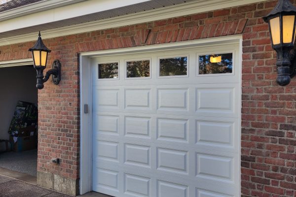 Traditional brick garage with a white raised-panel single door featuring four rectangular upper windows, flanked by black and gold lantern-style wall sconces and a paved driveway.