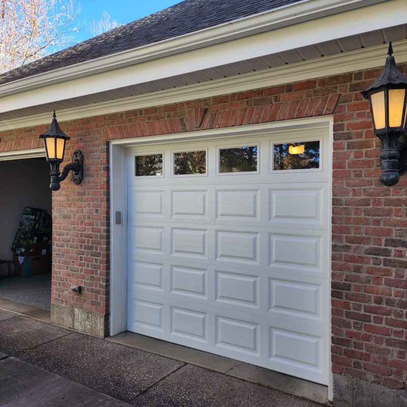 Traditional brick garage with a white raised-panel single door featuring four rectangular upper windows, flanked by black and gold lantern-style wall sconces and a paved driveway.
