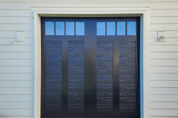 Single dark brown carriage-style garage door with upper window inserts, framed by cream-colored horizontal siding and white trim, set against a clean lawn and concrete walkway.