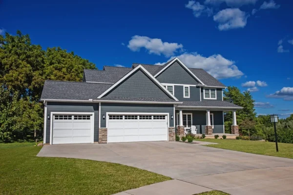 Spacious two-story home with blue-gray siding and two white paneled garage doors featuring upper windows, accented by stone bases and set on a large concrete driveway with a manicured lawn.