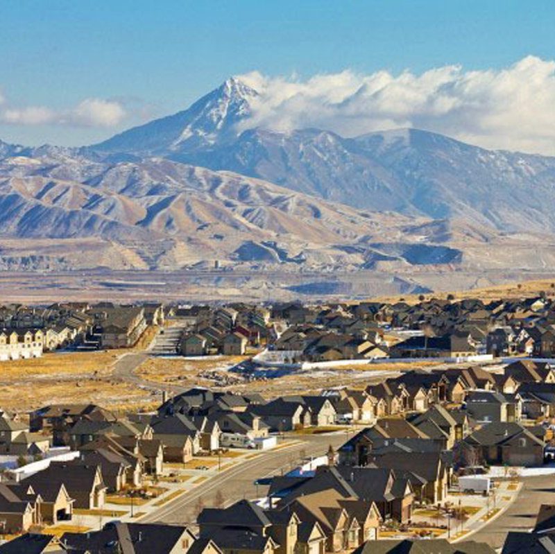 Expansive suburban housing development with rows of rooftops stretching toward rugged, dry mountains and a prominent peak under a partly cloudy sky.