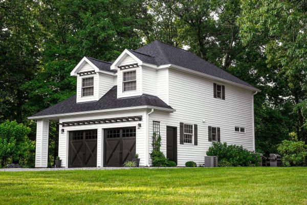 Charming two-story white house with black carriage-style garage doors featuring X-brace details and small upper windows, surrounded by lush greenery and set against a wooded backdrop.