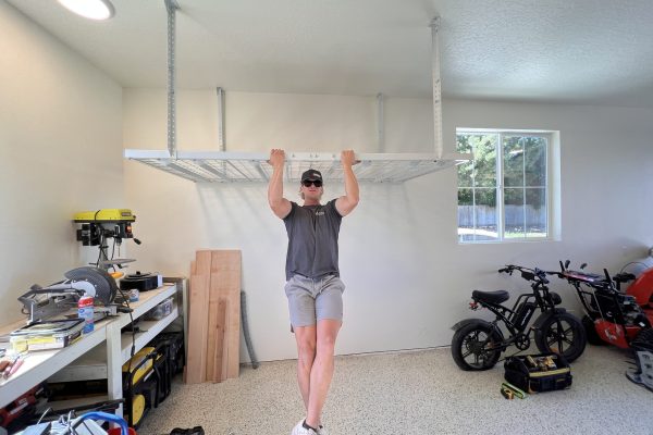 Man performing a pull-up on a ceiling-mounted garage storage rack, showcasing its strength and stability, in a well-organized garage with tools and electric bikes.