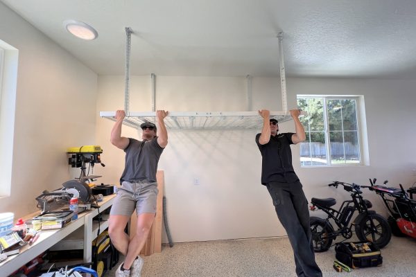 Two men performing pull-ups on a ceiling-mounted garage storage rack to demonstrate its strength, surrounded by tools, electric bikes, and natural light from nearby windows.